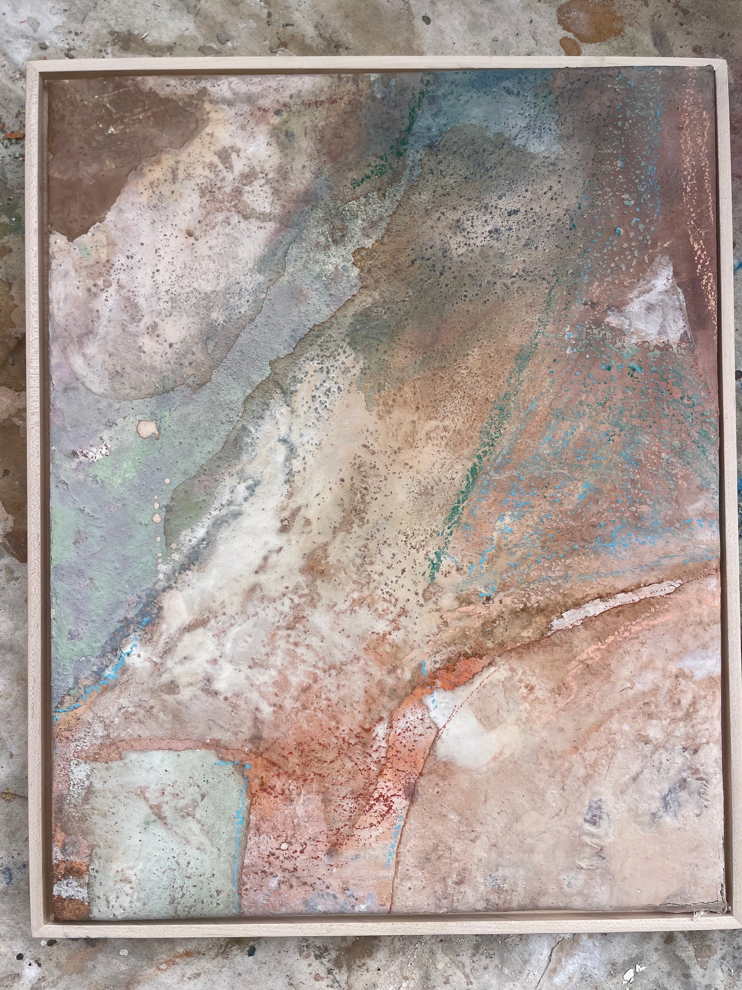 An abstract mixed-media painting in a square wooden frame, featuring textured washes of beige, brown, muted blue, and coppery orange tones on a concrete floor.